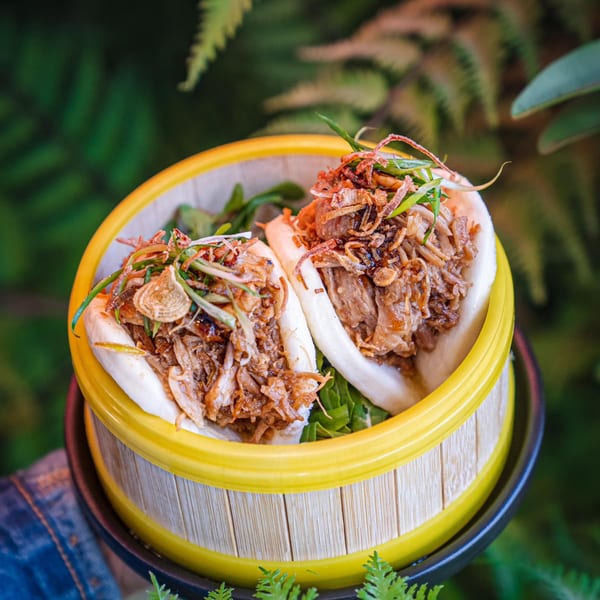 Two NōKA Ramen & Bar pork belly bao buns with crispy onions and scallions in a bamboo steamer.
