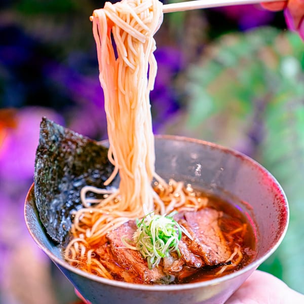 Chopsticks lift ramen noodles from a bowl with beef, green onions, and nori in a dark setting.