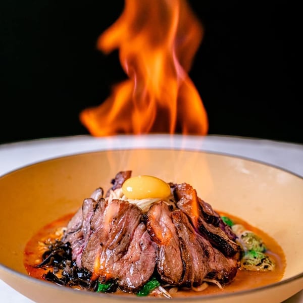 NōKA Ramen & Bar's ramen bowl with sliced meat, an egg yolk, and flames rising above.