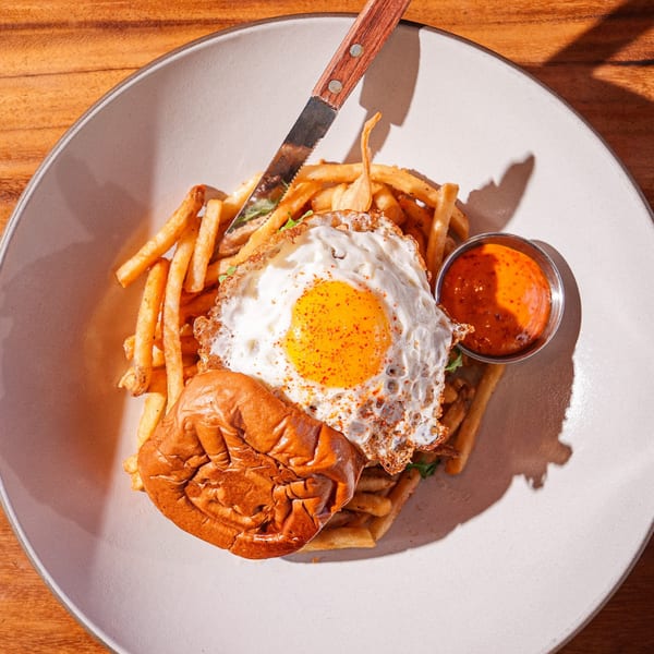 A burger with a fried egg and fries, served with a side of sauce on a white plate.